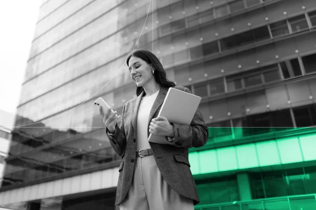 Business professional holding a laptop and checking her smartphone outside an office building