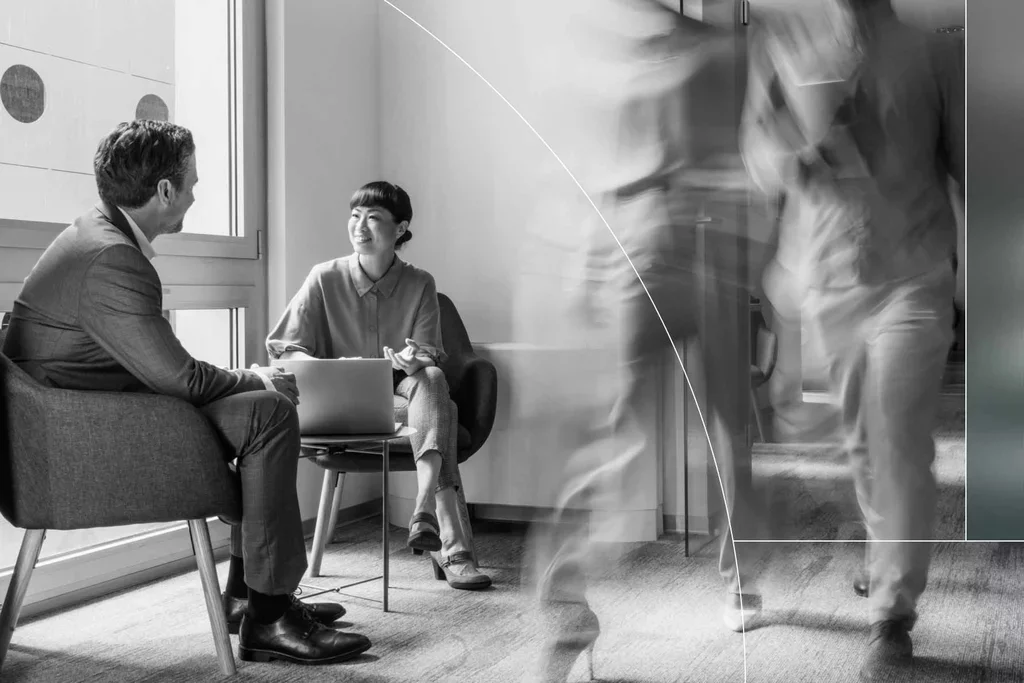 Two professionals discussing work in an office while colleagues walk nearby.