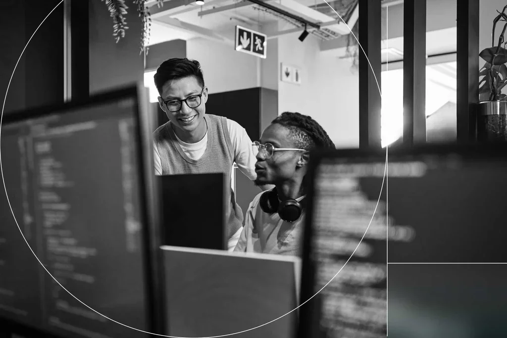 Two technology professionals collaborating at computer monitors in an office.