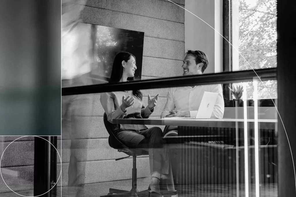 Two professionals discuss work together at a laptop in a modern office.