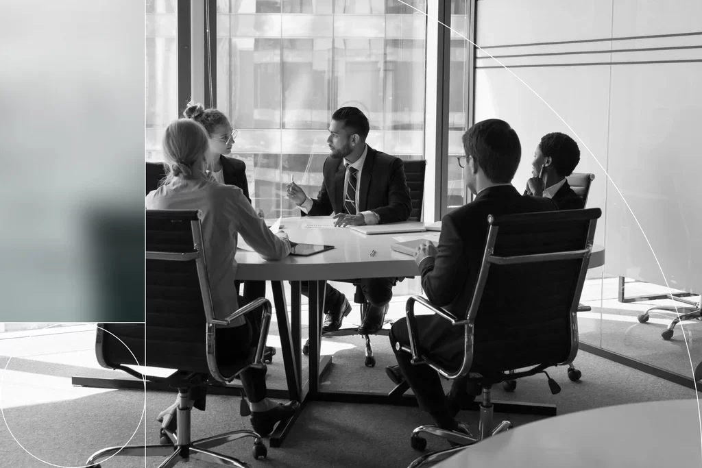 Five business professionals discussing documents during a meeting in a bright conference room.
