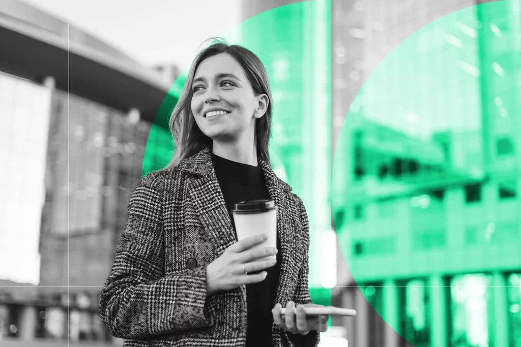 Business professional holding a smartphone and coffee while walking through a modern office.