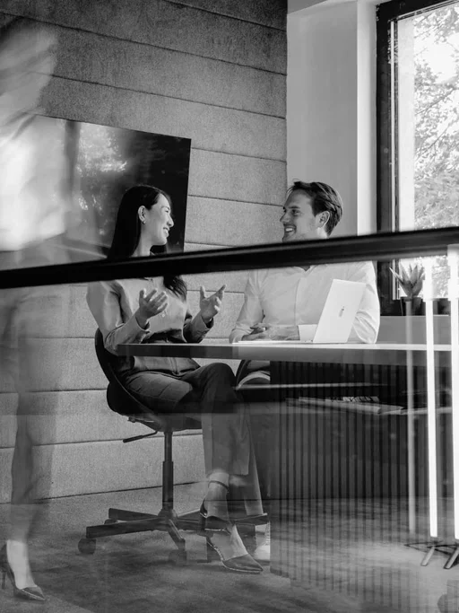 Two professionals discuss work at a table with a laptop in a modern office.