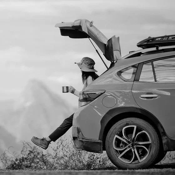 Person relaxing beside parked SUV with open hatch overlooking scenic mountain landscape.