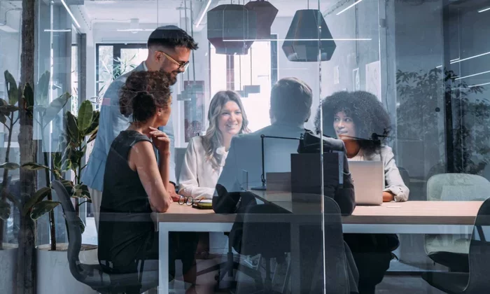A diverse team of security and IT leaders meeting in a glass conference room, collaborating around a table with laptops—symbolizing full accountability and governance across AI and enterprise security initiatives at Proofpoint.