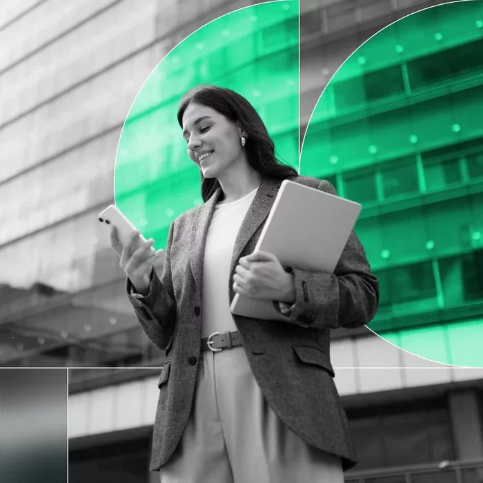 Business professional checks her smartphone while carrying a laptop outside an office building.