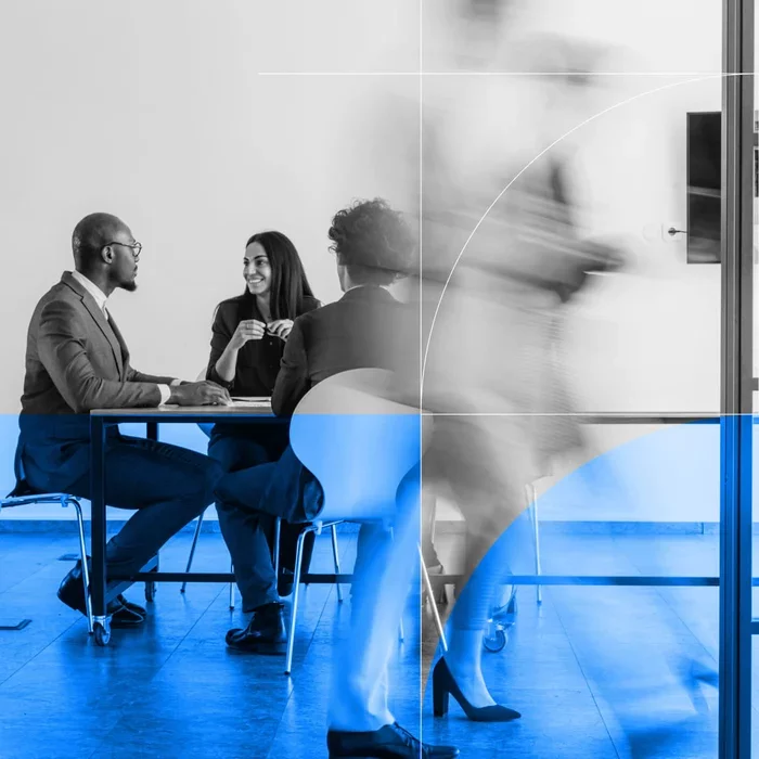 Three business professionals meeting at a table while a colleague walks past in the foreground.