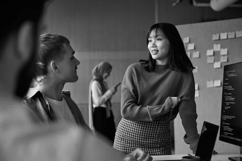 Two coworkers discussing information at a computer workstation in an office, with a third employee in the background.