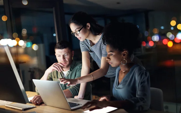 Three colleagues working together on laptop reviewing information at night.