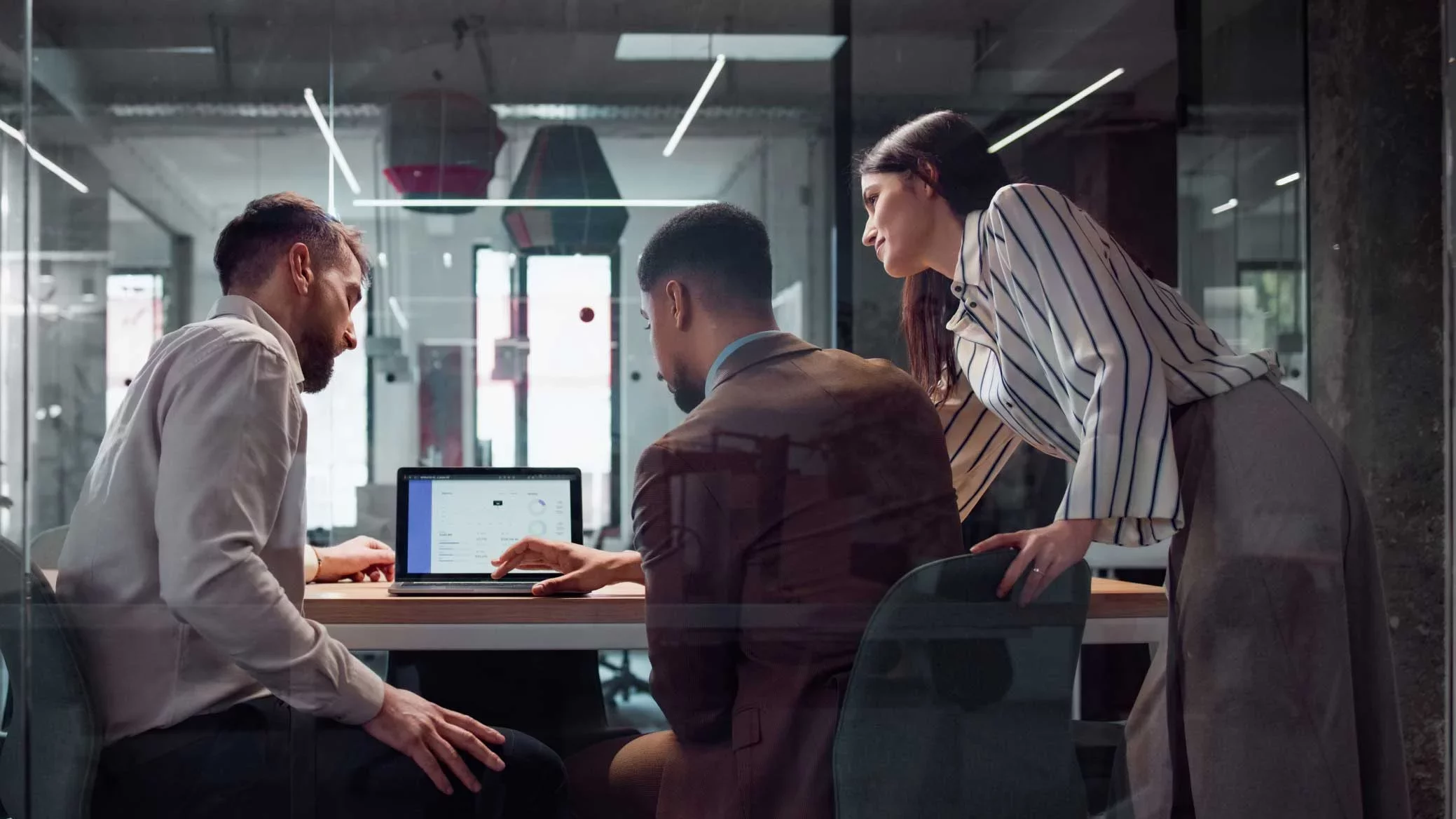 A cross-functional team collaborating around a laptop in a modern office, reviewing performance data—representing secure AI adoption at enterprise scale powered by Proofpoint.