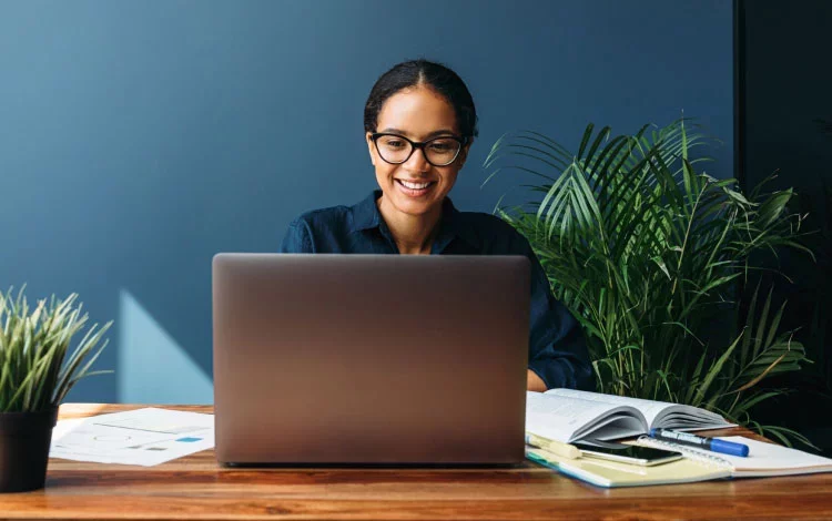 Woman using laptop at desk with documents and plants nearby.